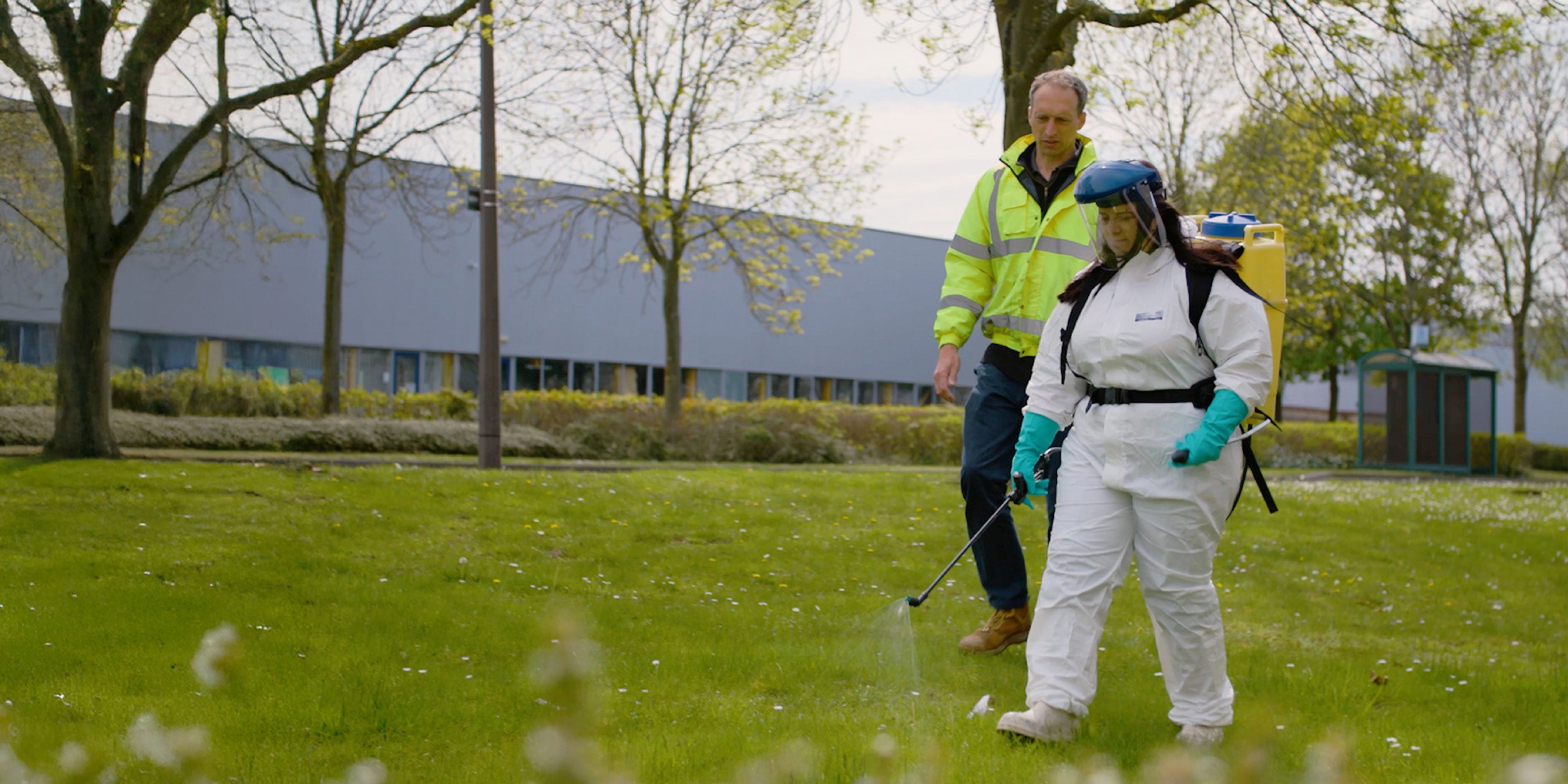 A woman wearing PPE being trained to use a sprayer by a man in high visibility clothing.