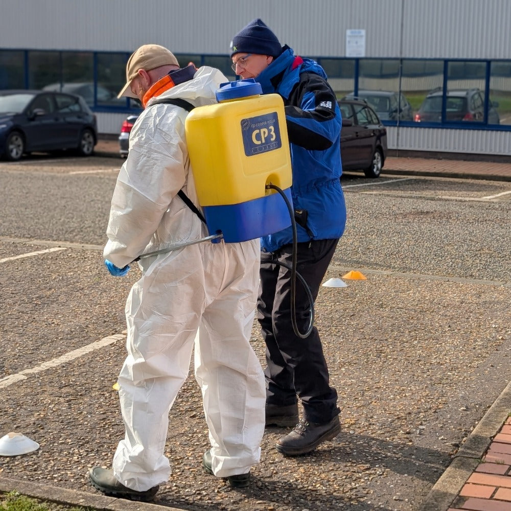 A person in a white hazmat suit wearing a yellow and blue CP3 backpack sprayer while another person in a blue winter jacket assists them in an outdoor paved parking lot.