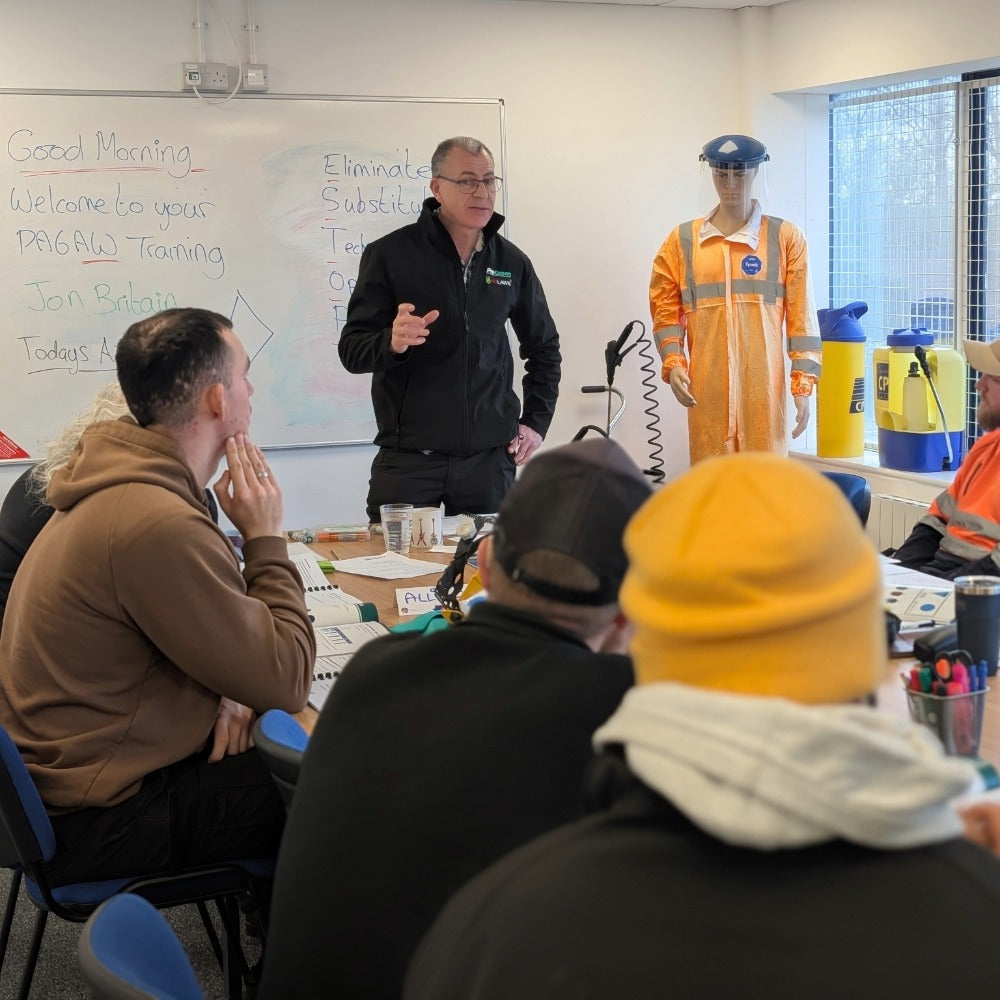 A PA6A/AW training session in progress, with the trainer in a black jacket speaking to a seated audience. A mannequin wearing a high-visibility orange work uniform stands near a window in the background, next to yellow containers.