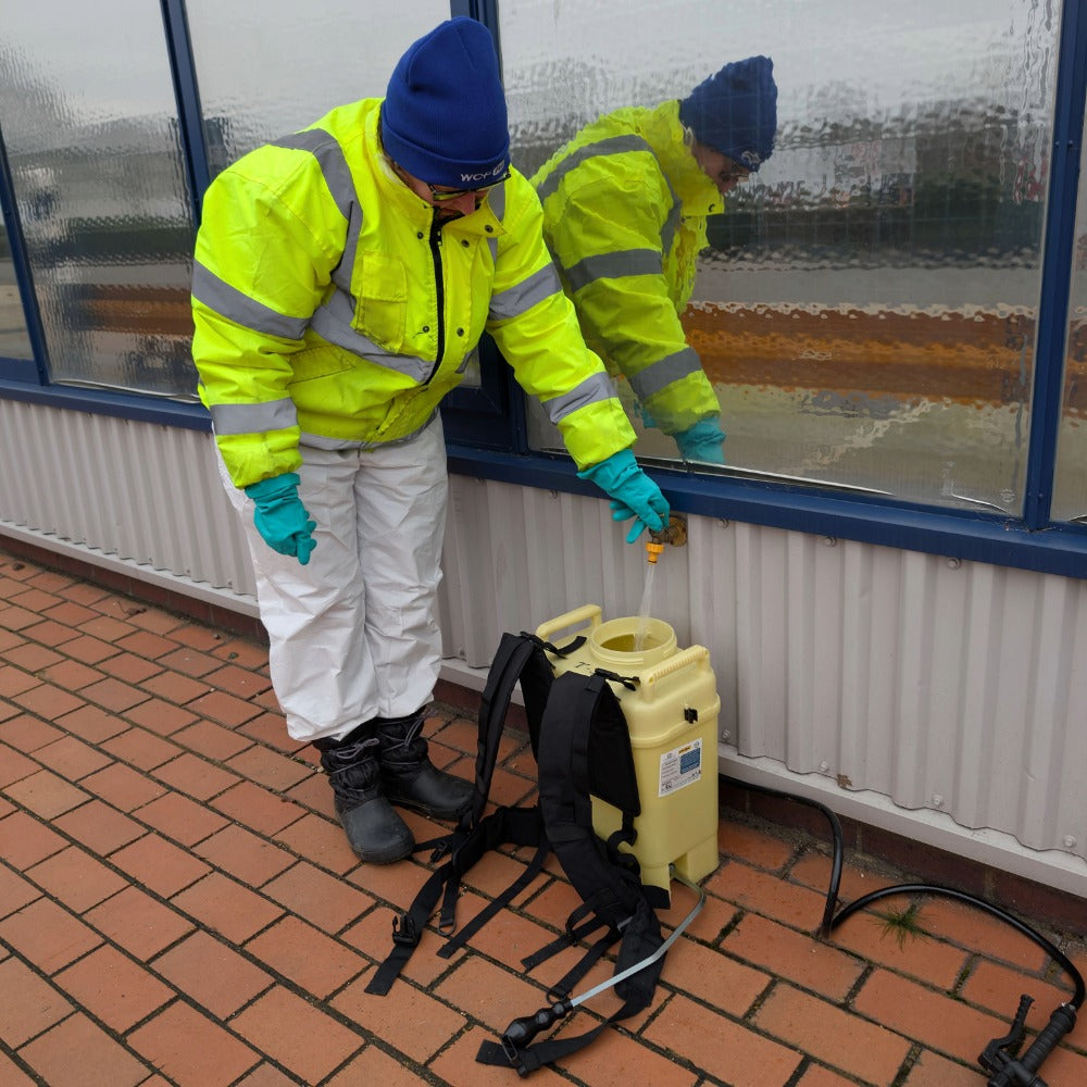 A person in a high-visibility yellow jacket, white coverall, protective footwear and gloves is filling up a yellow backpack sprayer on a brick patio beside a window.
